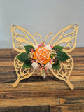 Papillon décoratif en bois ajouré, orné à la main de fleurs en cire parfumée orange, blanches et roses avec feuillage vert.