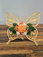 Papillon décoratif en bois ajouré, orné à la main de fleurs en cire parfumée orange, blanches et roses avec feuillage vert.